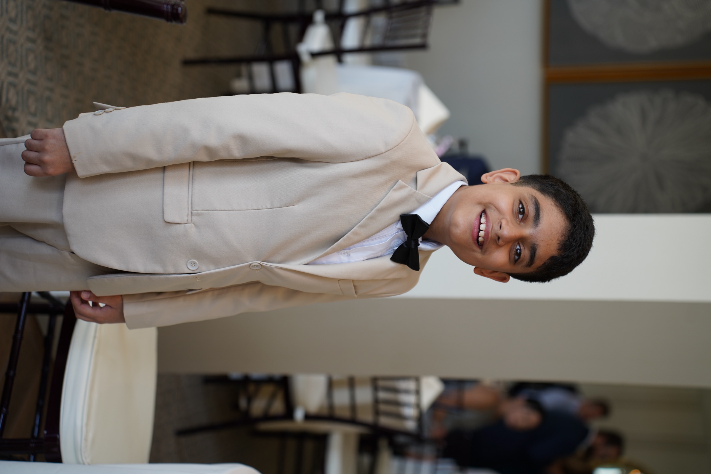 Boy in a tan suit and bow tie grinning at a wedding reception