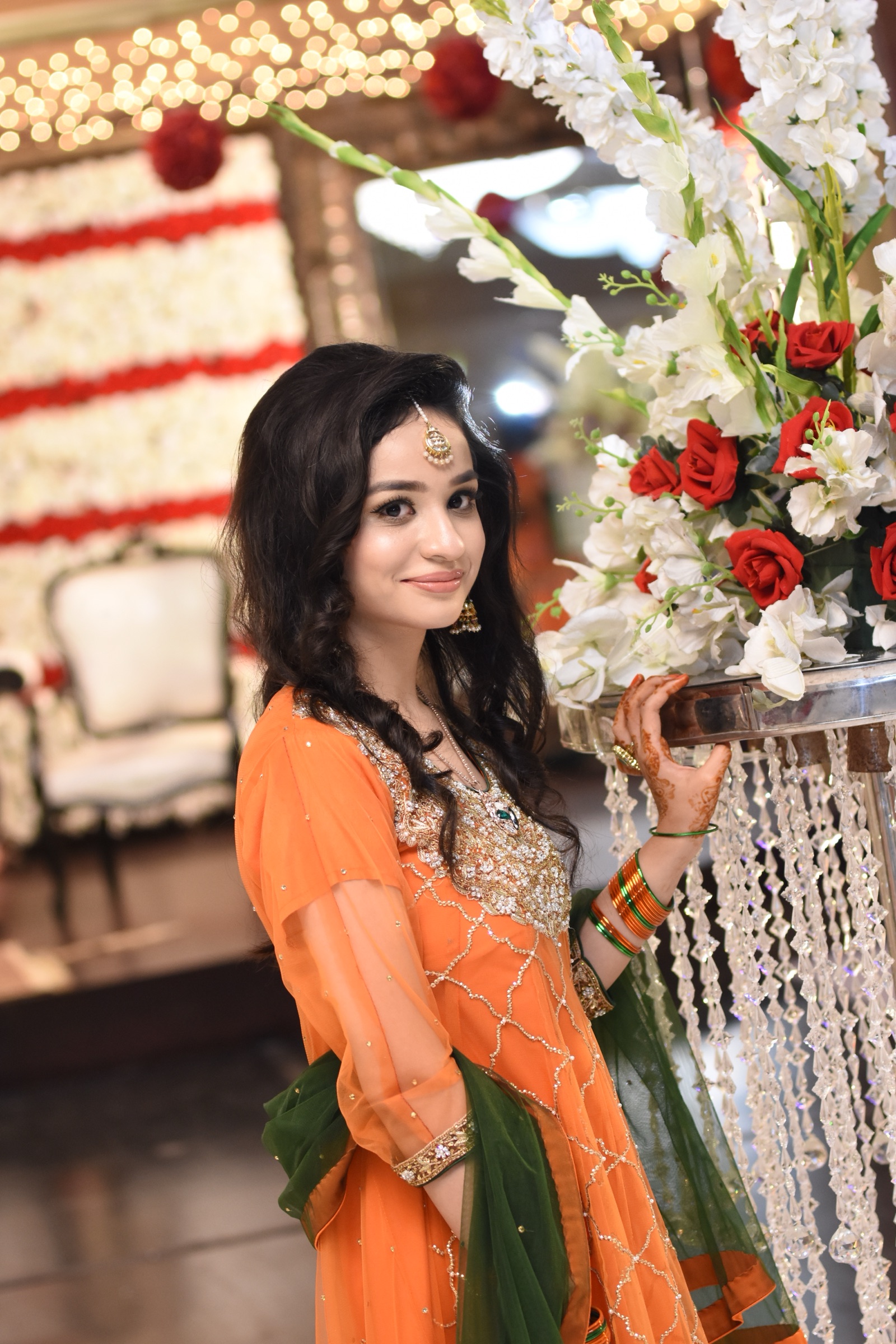 Guest in an embroidered orange dress beside the flower arrangements at a mehndi celebration